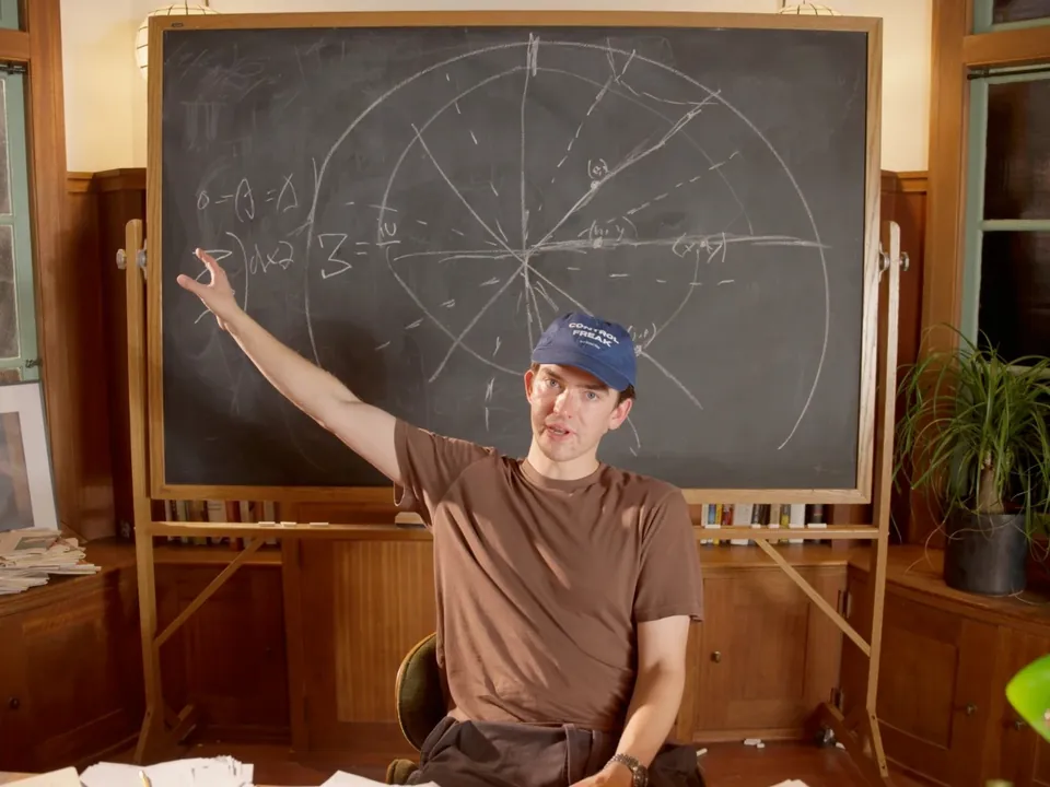 Nicholas in a cap pointing at chalkboard diagrams in the wood-paneled study