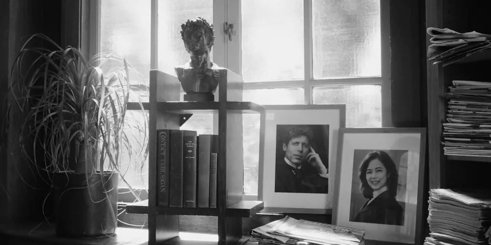 Black-and-white still of a bust, books and framed portraits on a shelf by a window